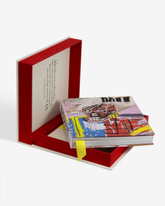 Basquiat: The World of Jean-Michel by Assouline: full product presentation — the open cream-and-red clamshell stands vertically in the background displaying the Basquiat quote on its ivory lid; in the foreground the book rests in the red tray, its colorful cover showing a Basquiat skull painting in vivid yellow, red, and blue with the word pneu; a yellow grosgrain ribbon bookmark trails from the binding