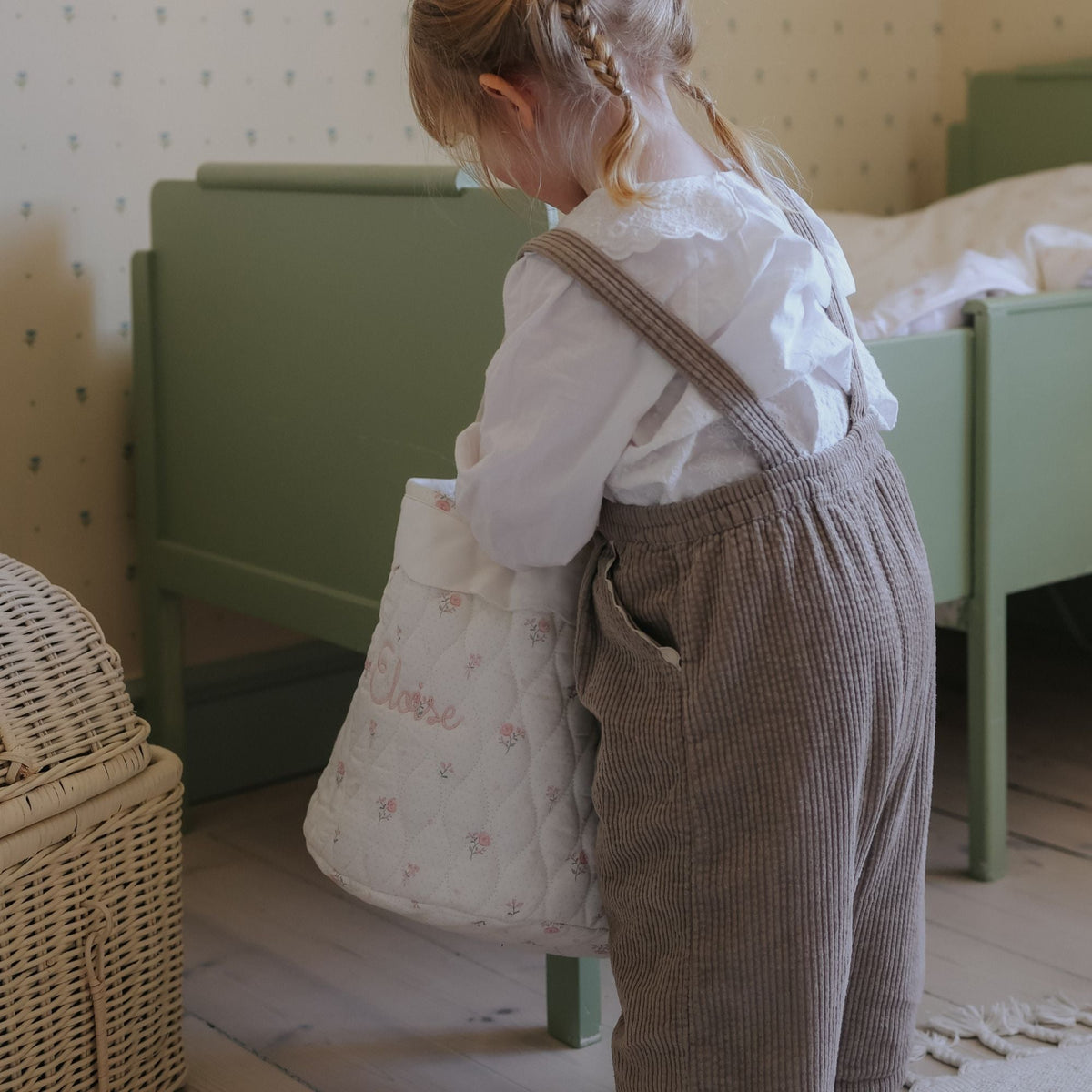 Child holding a patterned bag in a room with green furniture and a wicker basket.
