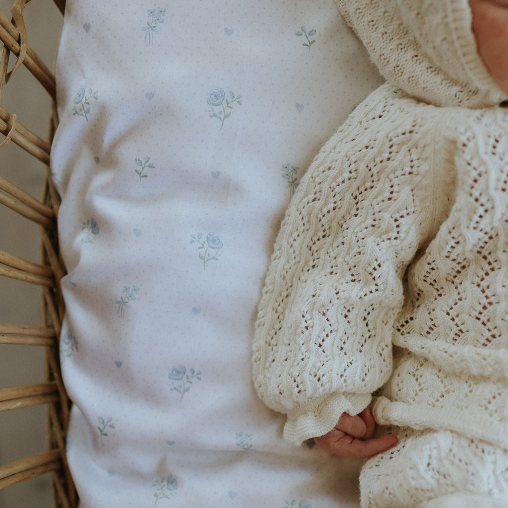 Baby wearing a white knitted sweater laying in a wicker crib with a floral-patterned crib sheet.