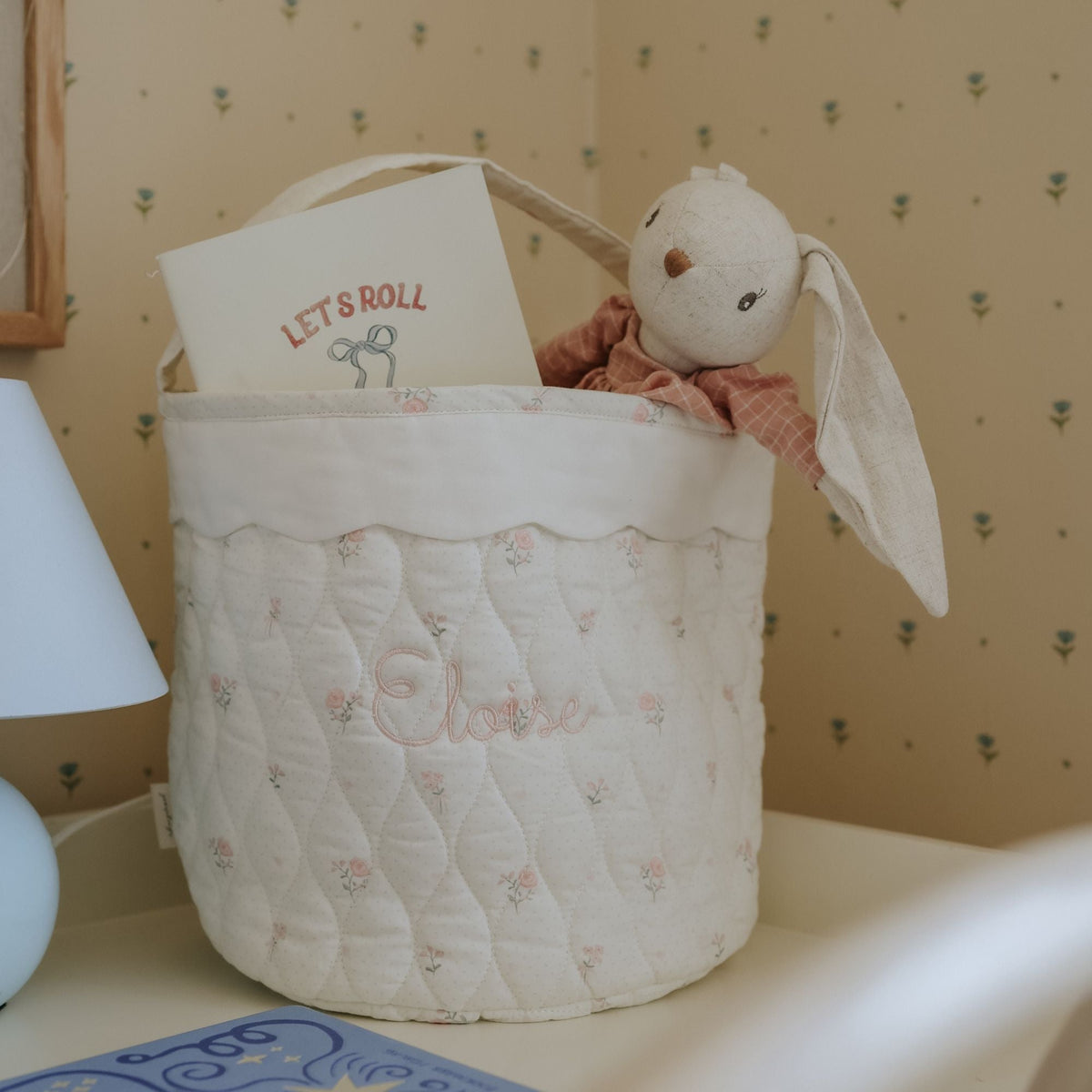 Quilted baby basket with plush toy and 'Let's Roll' book against a patterned wall.