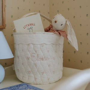 Quilted baby basket with plush toy and 'Let's Roll' book against a patterned wall.