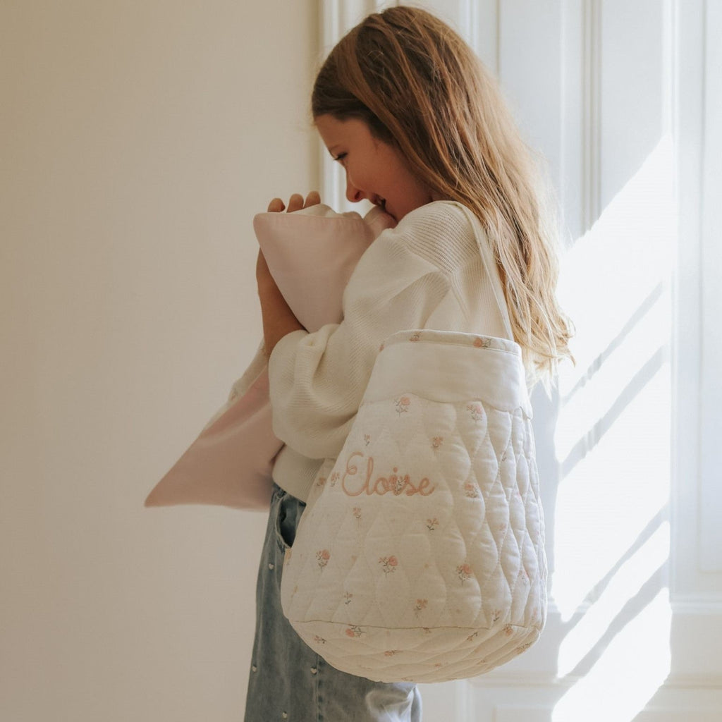 Child holding a baby wrapped in a quilt with 'Elise' embroidered on it, standing by a window.