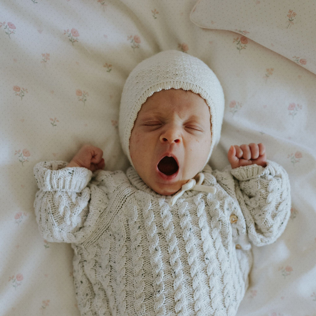 Newborn baby in a knitted outfit and hat yawning on a floral-patterned blanket.