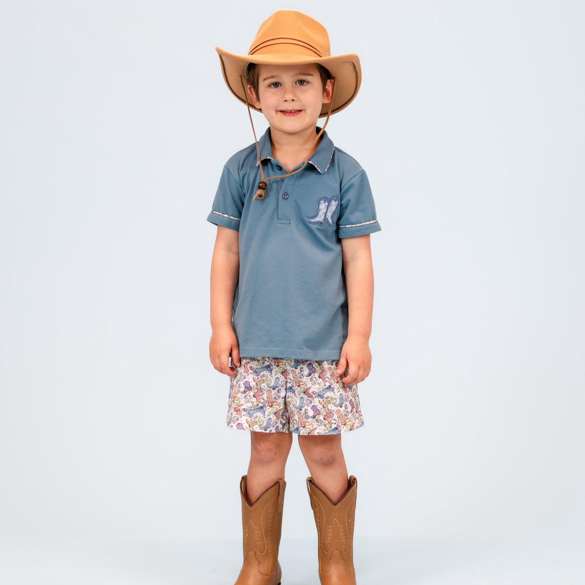 Front view of Fields boy polo set: blue polo with boot applique and floral trim, printed shorts, light gray background