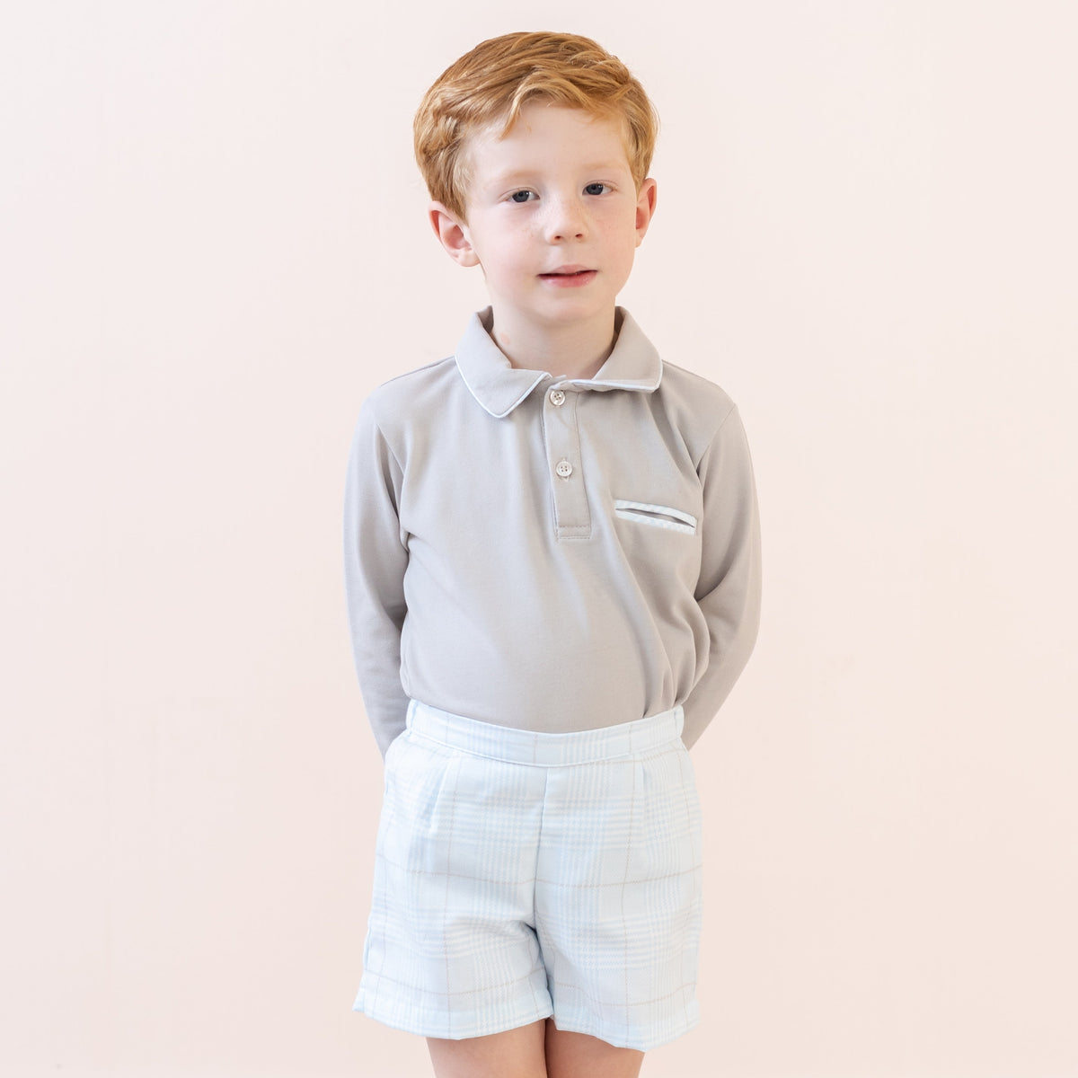 Front view of William boy set, beige long-sleeve polo with piped collar and chest pocket, light plaid shorts, pale pink background
