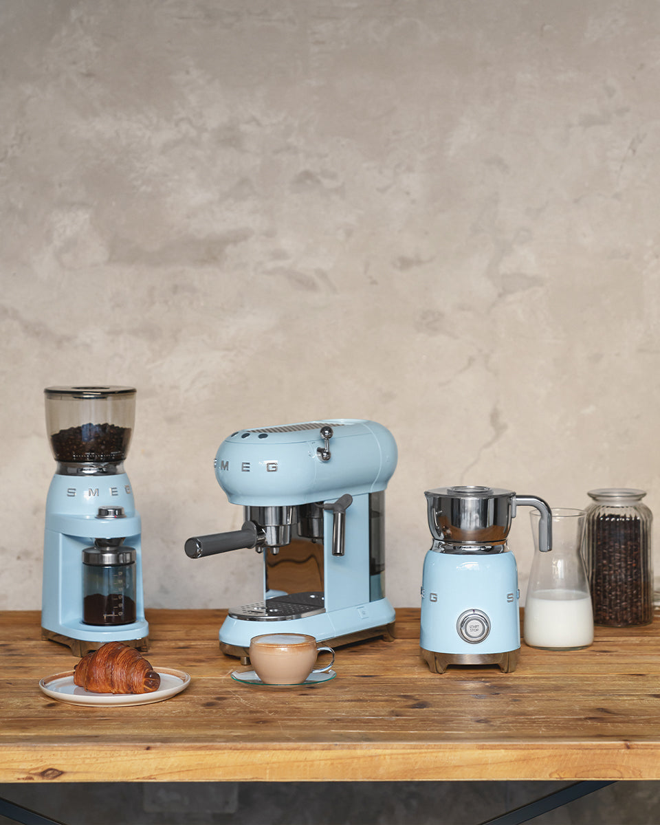 Front view of SMEG Milk Frother in Pastel Blue showcasing its stainless-steel jug on a wood counter with coffee appliances