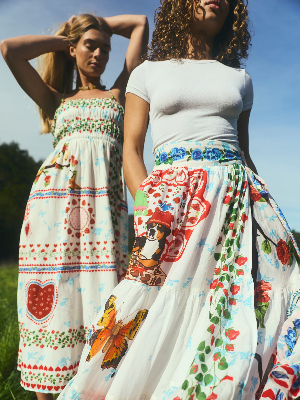 Two models wearing Alfie Wrap Skirts in Alfie White standing in a sunlit field showing colorful floral, butterfly, and bandana print