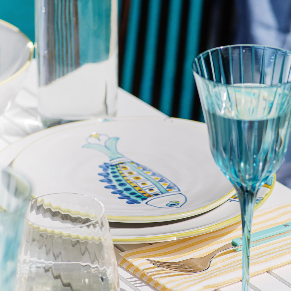 Angled view of Elba’s Fish waved-fin salad plate showing hand-painted blue and yellow fish motif with teal glassware