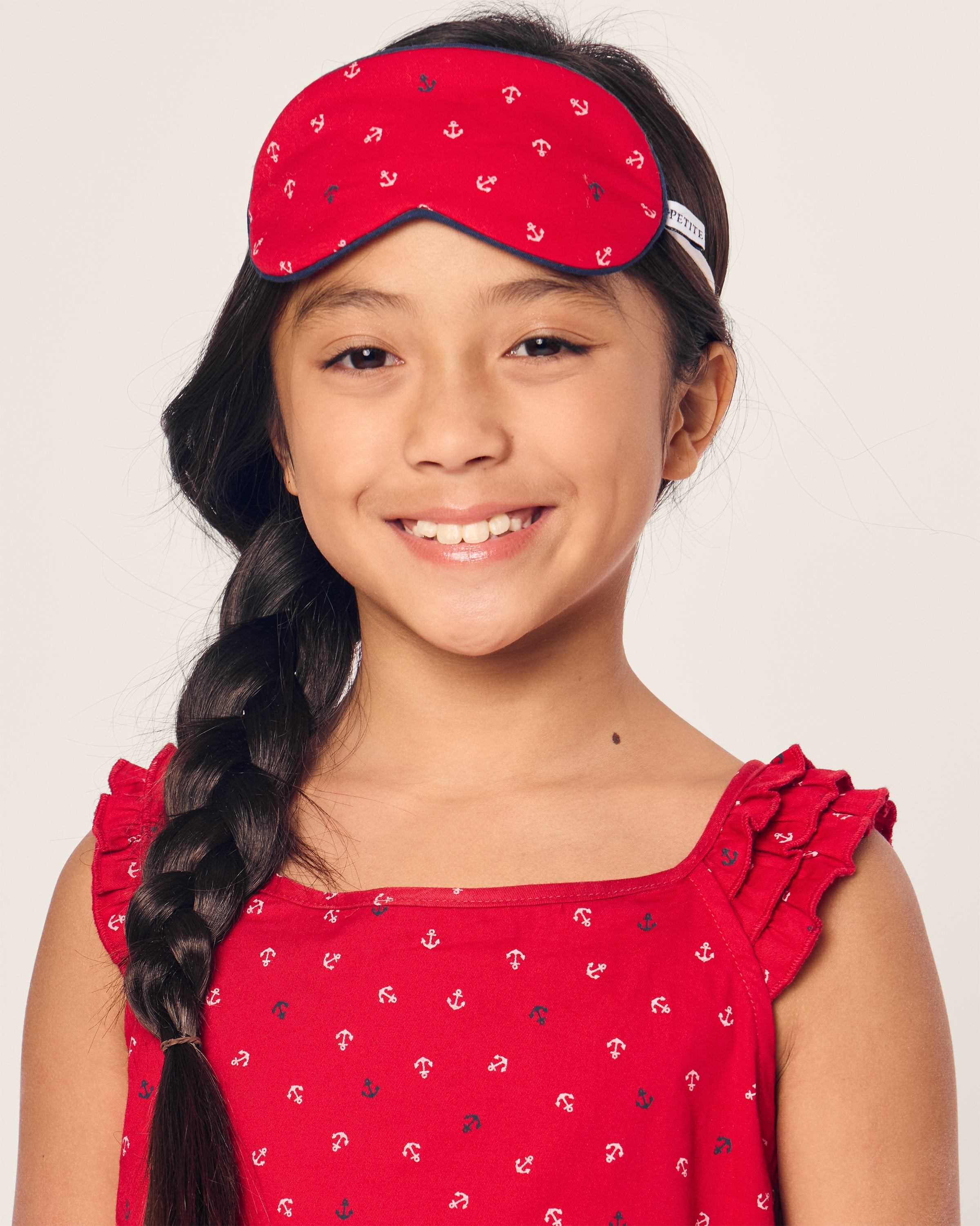 A young girl with long braided hair smiles at the camera, wearing Petite Plumes Kids Twill Sleep Mask in Anchors Away. She sports matching red pajamas with ruffled sleeves and a cute white anchor pattern, all set against a calming light-colored background.