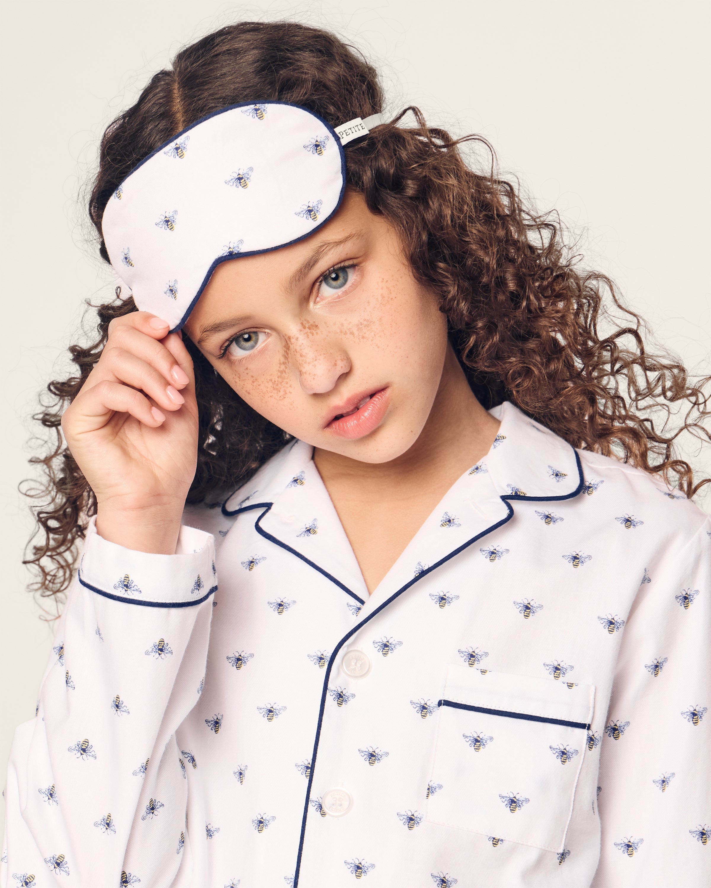 A young girl with curly hair wearing white pajamas decorated with buzzing bees adjusts her Petite Plume Kids Twill Sleep Mask in Buzzing Bees, gazing neutrally at the camera against a light background.