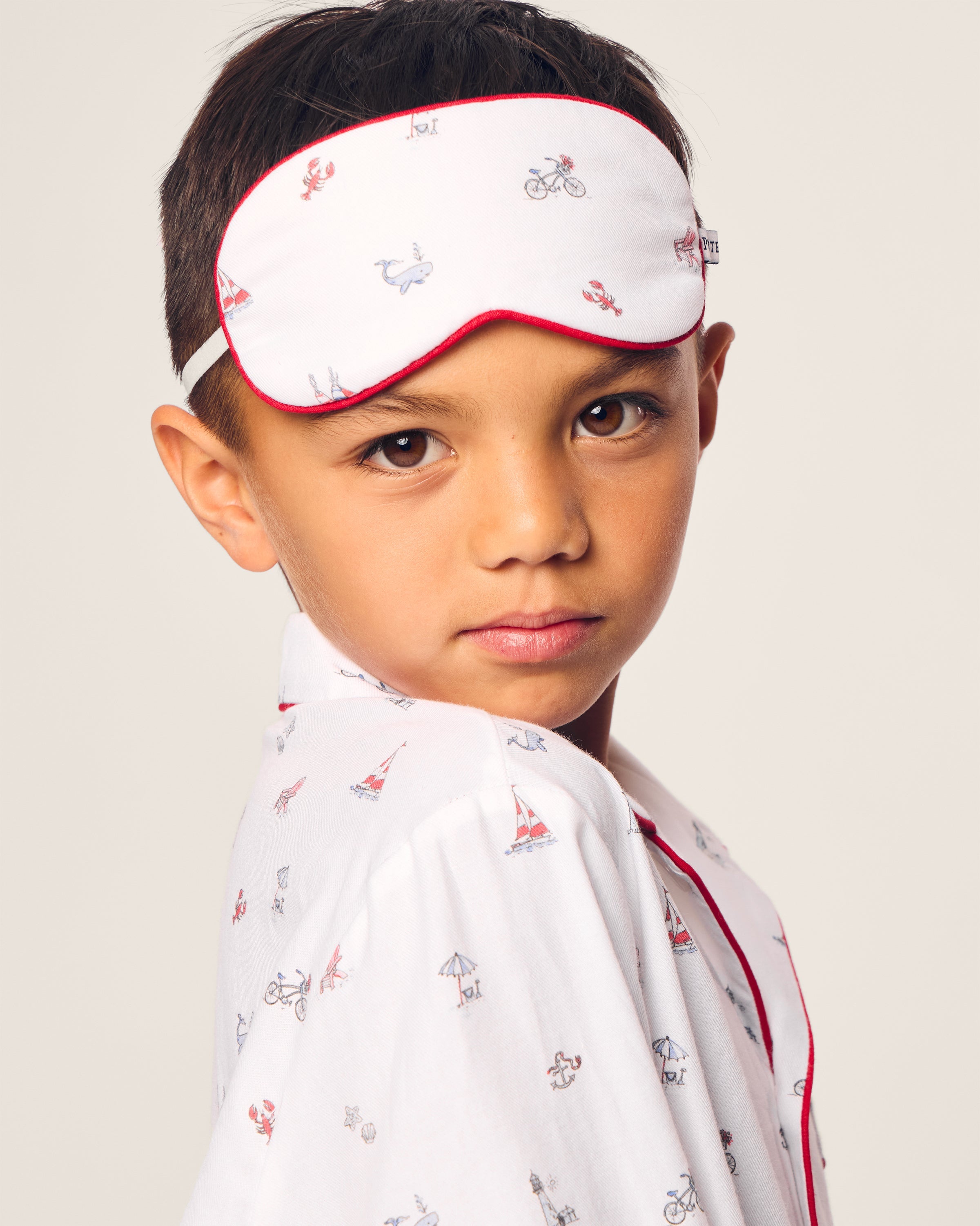 A child drifts into dreamland in white pajamas with small printed designs and red piping, wearing the Petite Plumes Kids Coastal Charms Eye Mask monogrammed as a gift, their expression neutral against a plain background.