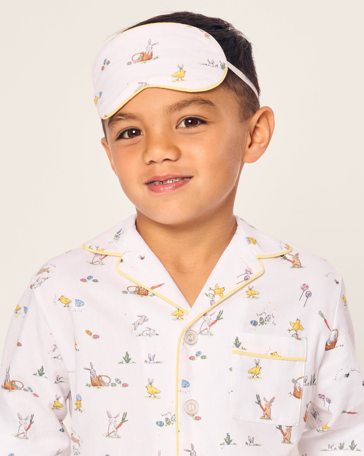 A young boy wears a white pajama set with animal and nature patterns, paired with a Petite Plume Kids Twill Sleep Mask in Easter Frolic on his forehead. He stands against a plain background, smiling gently.