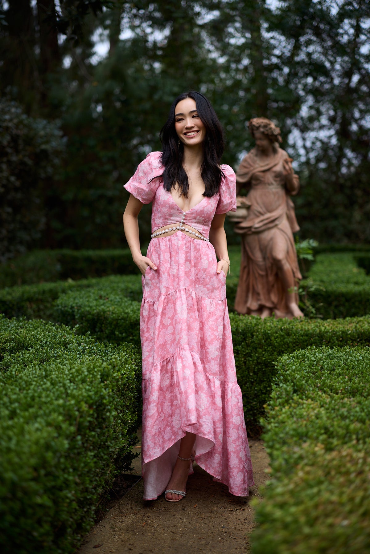Front view of Emma Dress in pink peony print with puff sleeves, V-neck and pearl cutout, asymmetric hem and pockets, garden setting
