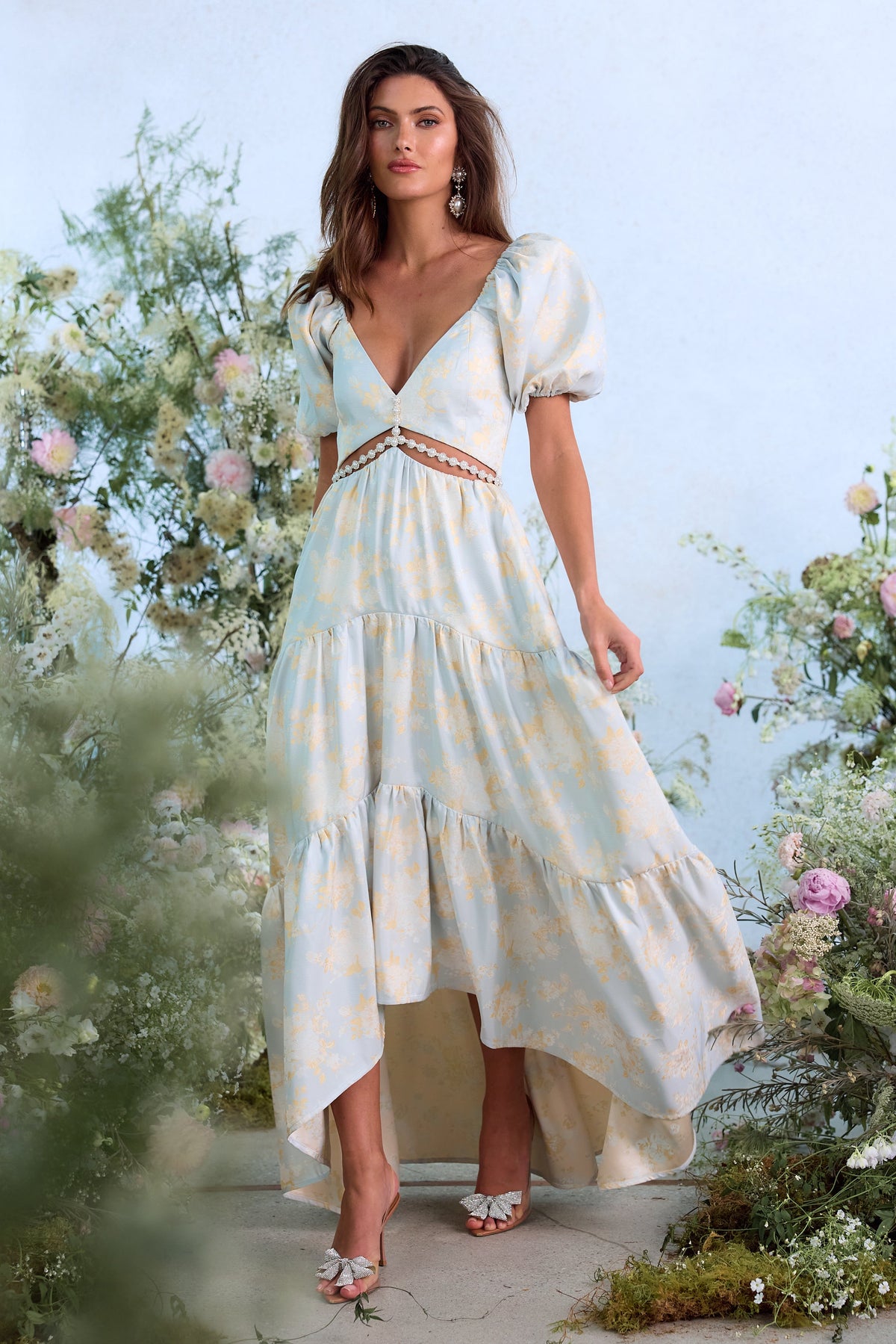 Front view of The Emma Dress in Sky Windsor Brocade showcasing cutout bodice and poof sleeves in a flower garden