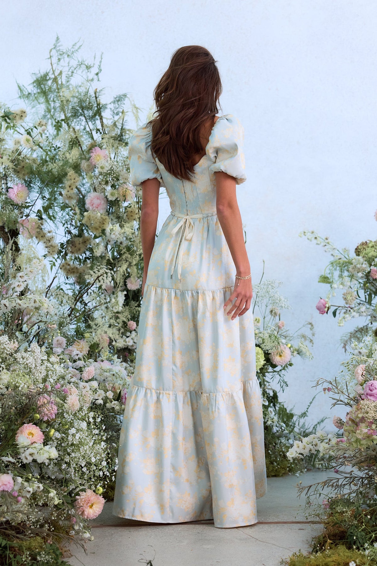Back view of The Emma Dress in Sky Windsor Brocade showcasing puff sleeves and gathered tiers in a flower garden