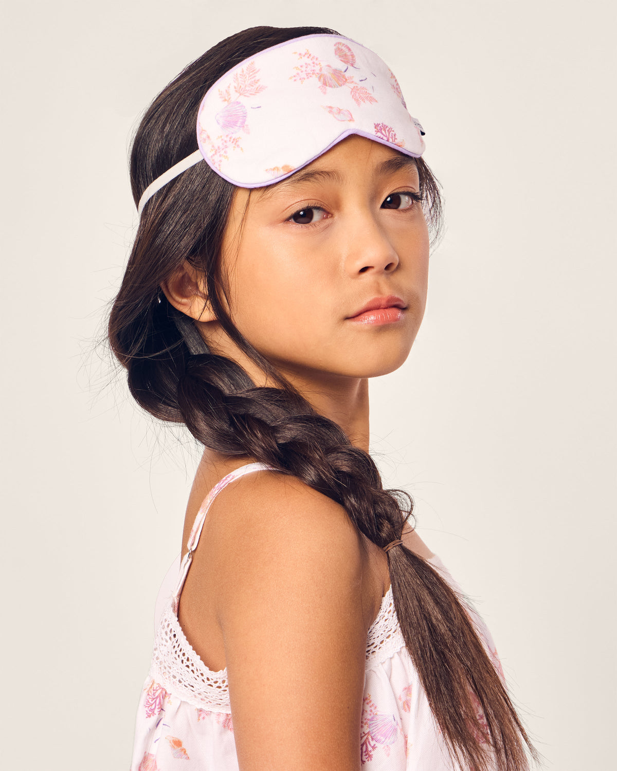 A young girl with long braided hair wears Petite Plumes Kids Twill Sleep Mask in Seashell Splendor, matching her light pink floral top. The eye mask rests on her forehead as she gazes to the side against a plain background.