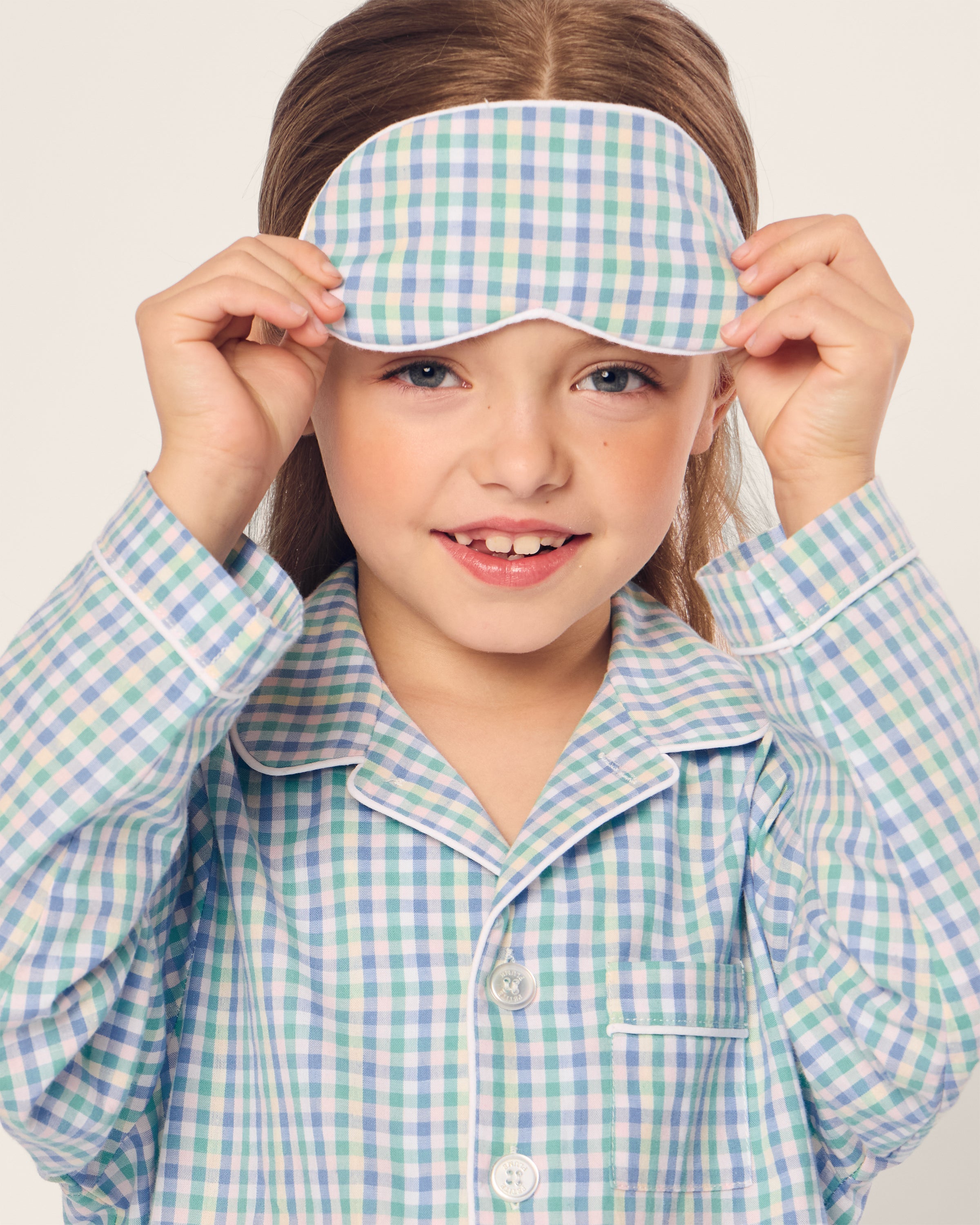 A young girl with long hair dons a Kids Twill Sleep Mask in Spring Gingham by Petite Plume along with matching pastel checkered pajamas. Her smile beams against a plain background, radiating the charm of a perfectly monogrammed gift.