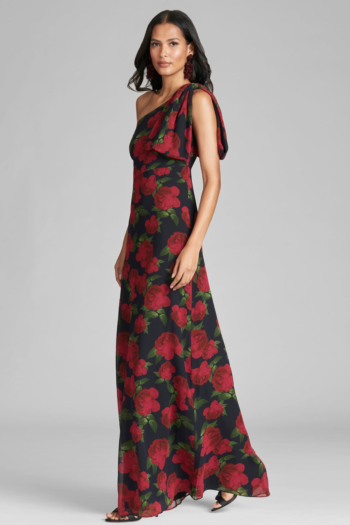 Three-quarter view of Chelsea Gown in Ruby Tea Roses showcasing one-shoulder bow and floral chiffon on gray background