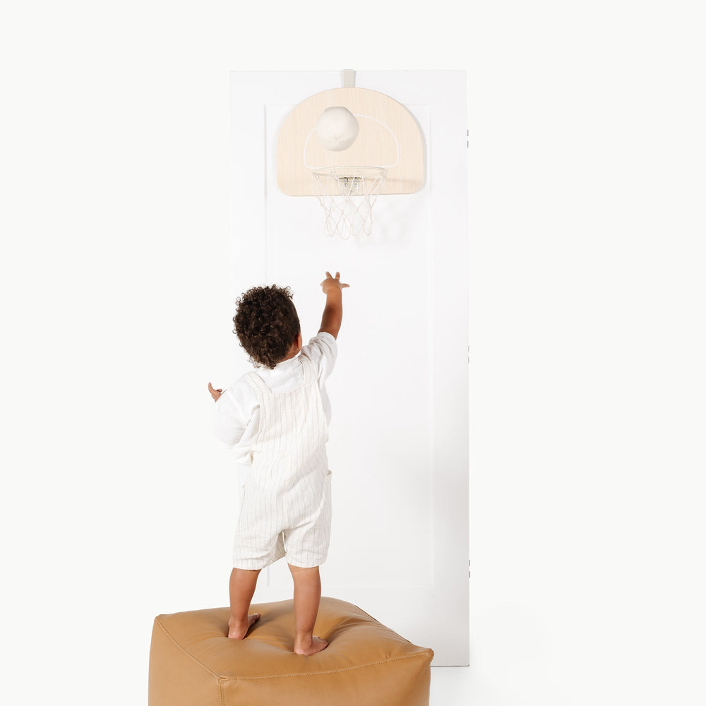 Front view of doorway basketball hoop on white door showcasing light wood backboard and spring rim with toddler shooting from tan pouf