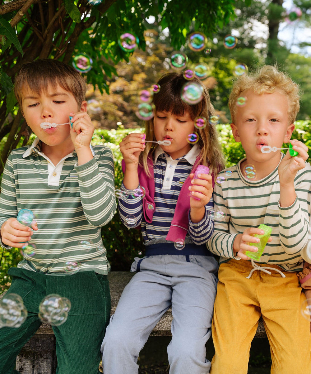 Three children sitting on a bench outdoors blowing bubbles, two wearing Edward T-Shirt in Pine Stripe
