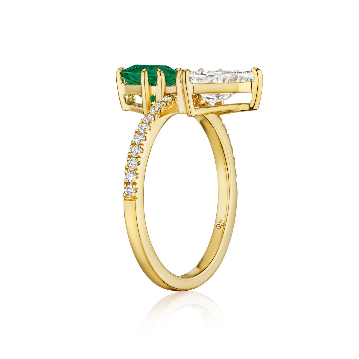 The Emerald and Diamond Double Shield Ring in Yellow Gold features a diamond band and two emerald-cut gems—one green, one white—set side by side in elegant claw settings.
