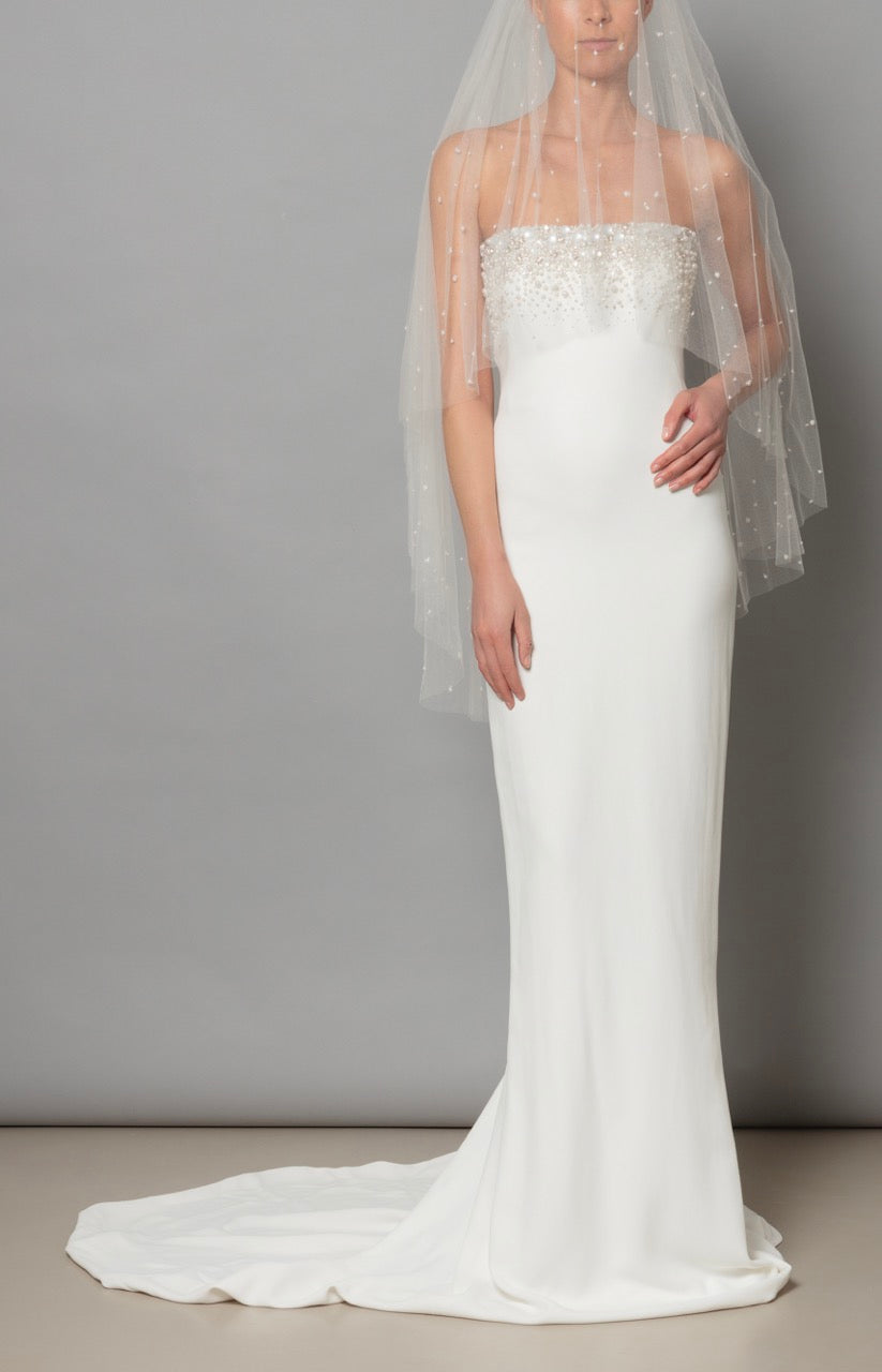 Front view of the Eve Dress in Ivory showcasing a strapless, slim fit, beaded bodice, veil, and train on gray background