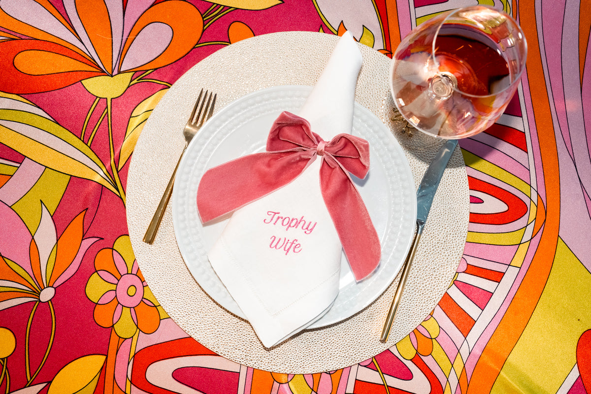 Top view of white linen napkin embroidered Trophy Wife, tied with a pink velvet bow on a plate with gold flatware