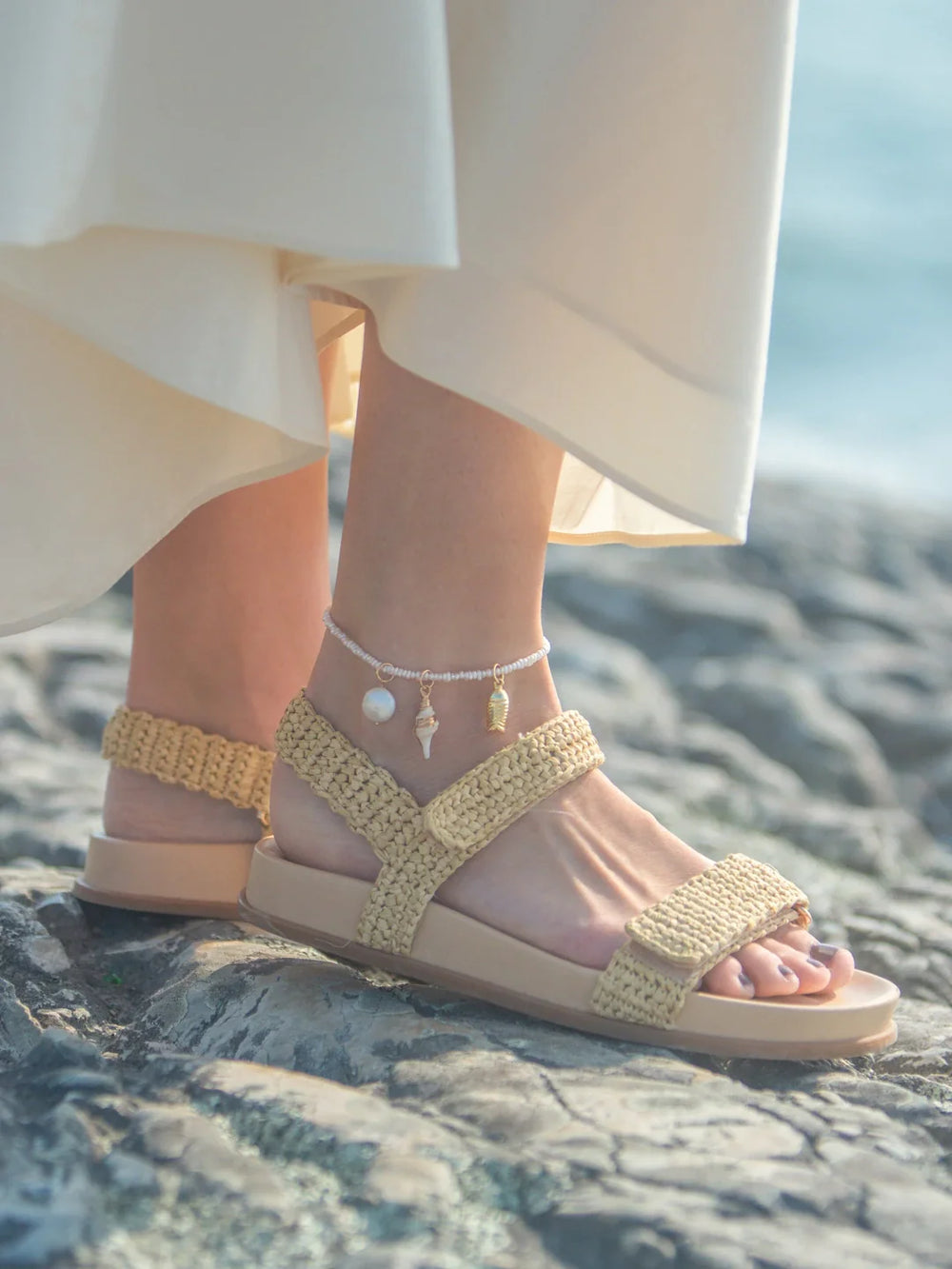 Close-up of Finn Anklet on model ankle showcasing freshwater pearls and gold shell charms on a rocky beach