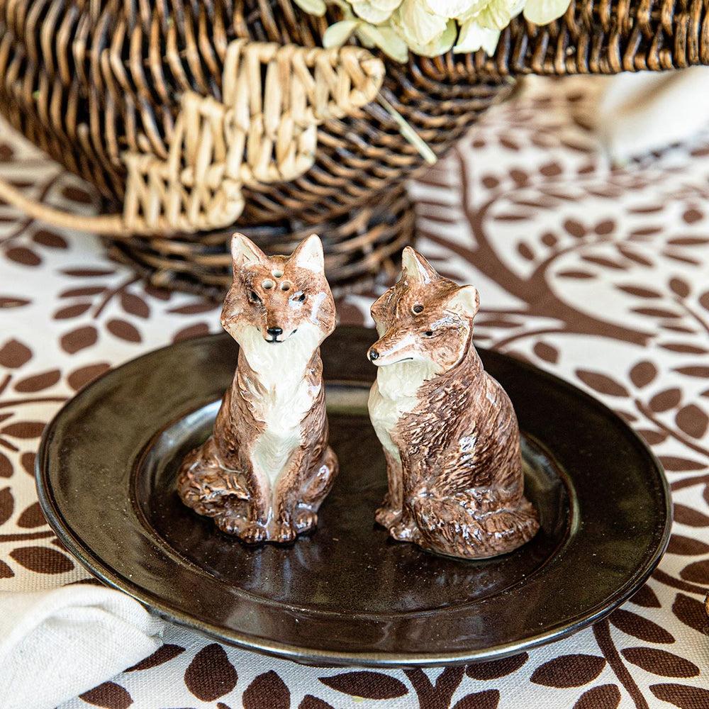 Front view of Clever Creatures fox salt and pepper set showcasing two glazed ceramic fox shakers with walnut finish on a dark plate and woven basket background