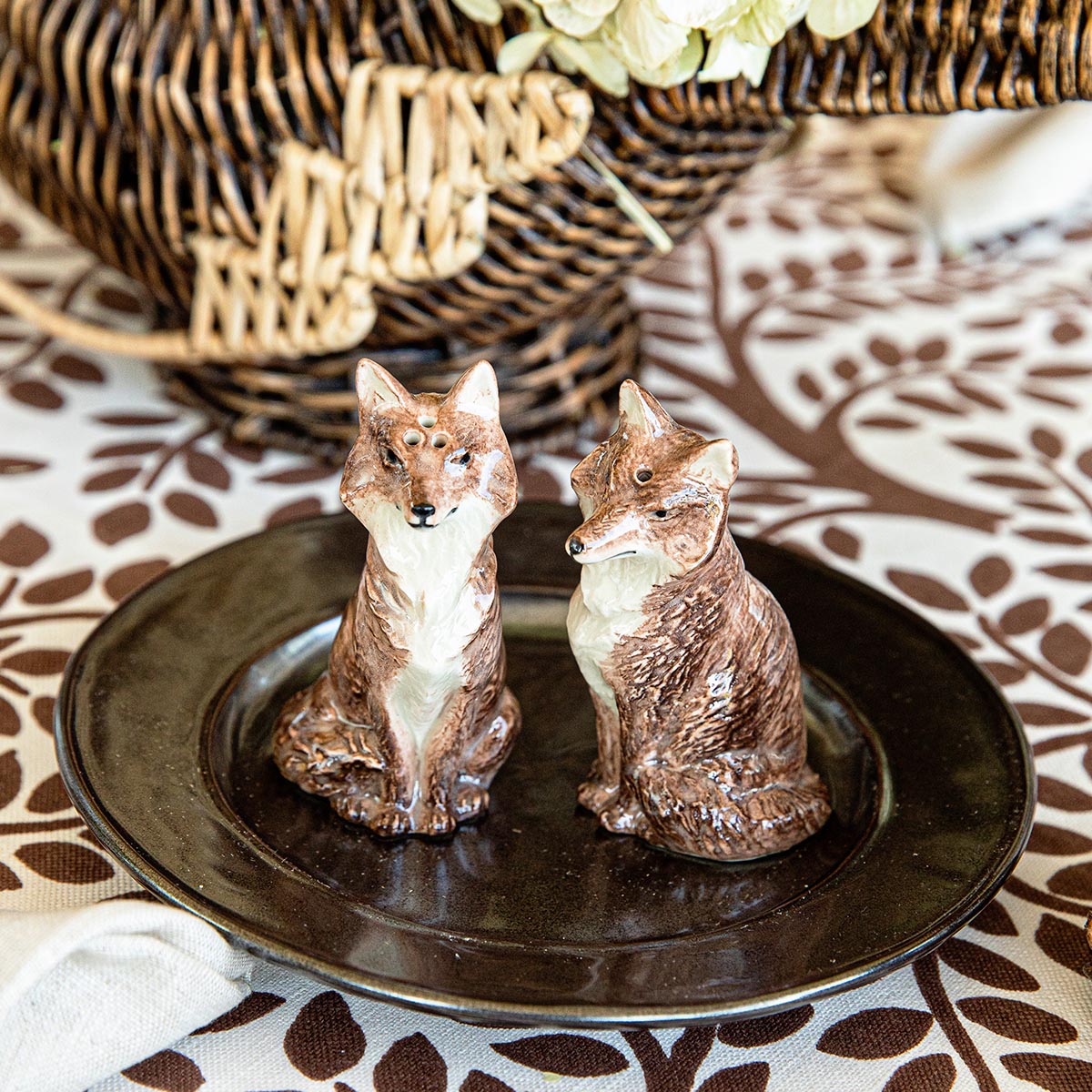 Front view of Clever Creatures fox salt and pepper set showcasing two glazed ceramic fox shakers with walnut finish on a dark plate and woven basket background