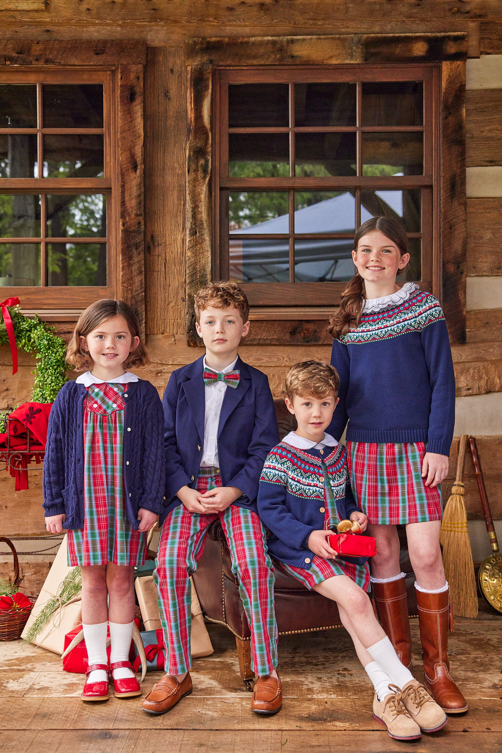 Four children wearing navy Highlands Fair Isle cardigans and tartan outfits posed in front of a rustic wooden cabin
