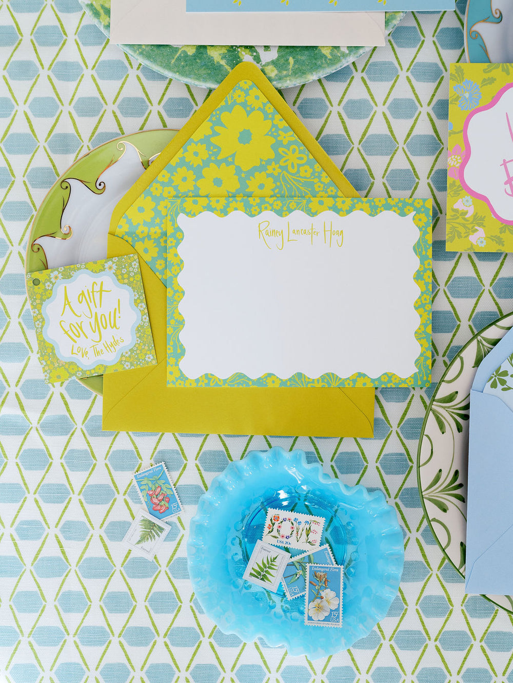 Top-down view of Chartreuse Ditsy Floral stationery set showcasing scalloped notecard with personalized gold script and floral-lined envelope on blue-green geometric tablecloth