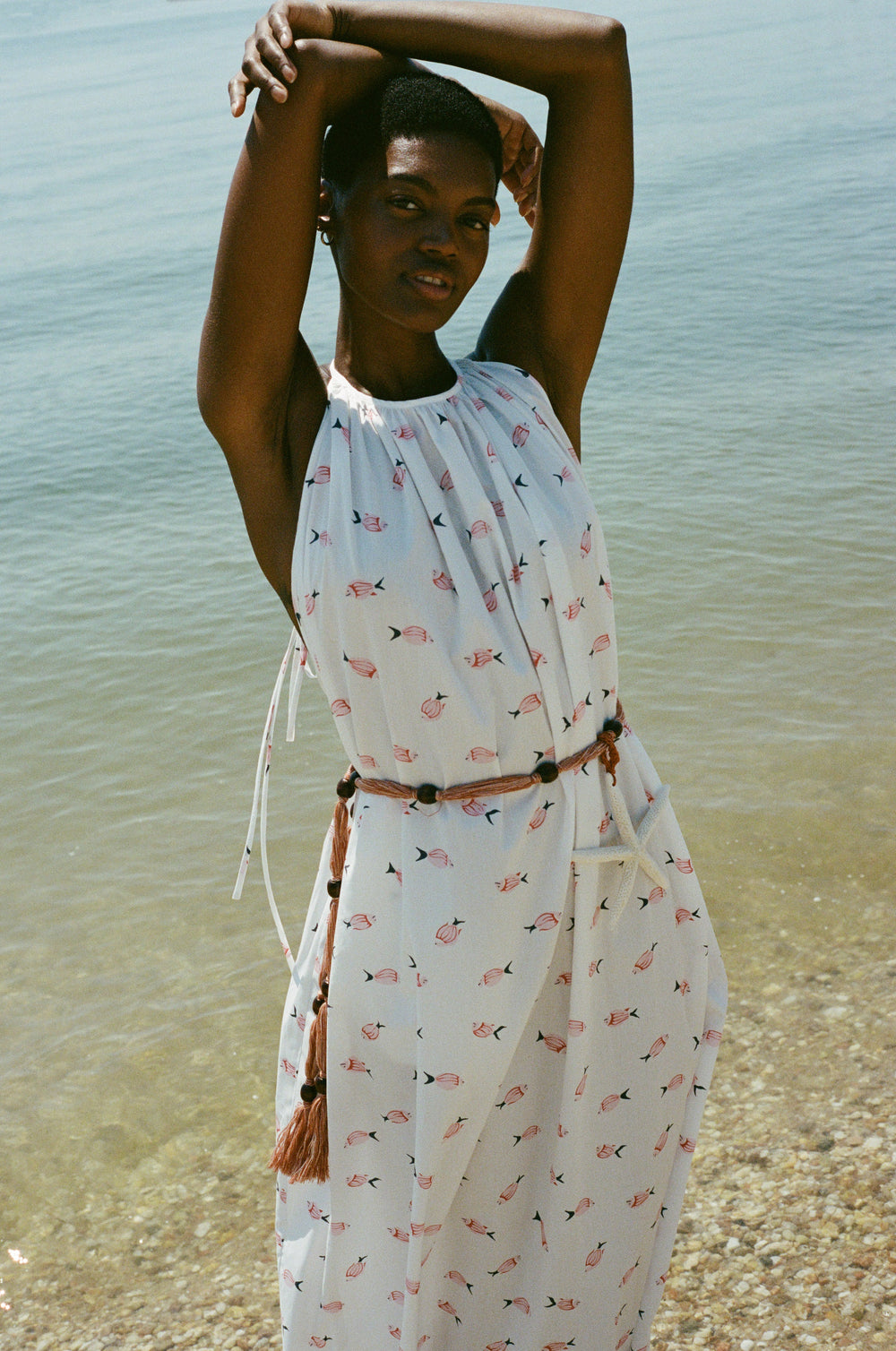Model wearing halter dress in vintage pink fish print with gathered neckline, braided belt, ankle-length, at water’s edge