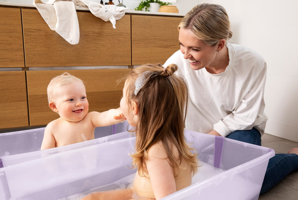 Two young children playing in side-by-side lavender Flexi Bath extra-large tubs while a smiling mother watches in a bright bathroom