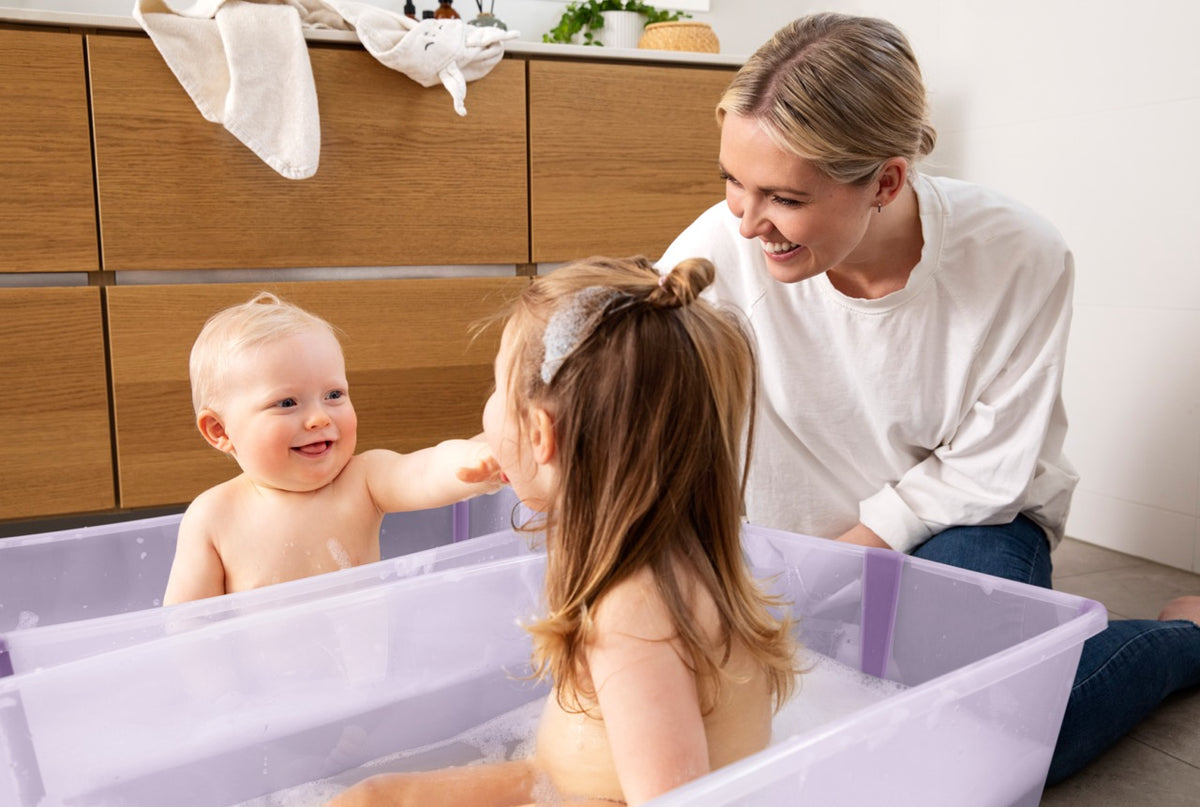Two young children playing in side-by-side lavender Flexi Bath extra-large tubs while a smiling mother watches in a bright bathroom