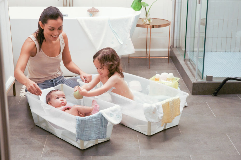 Mother kneeling beside two young children bathing in white Flexi Bath X-Large tubs on a tiled bathroom floor