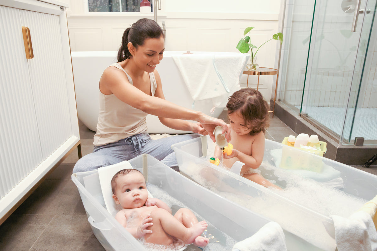 Mother bathing a toddler and an infant in two white Flexi Bath X-Large tubs on a bathroom floor, with bubbles and bath toys