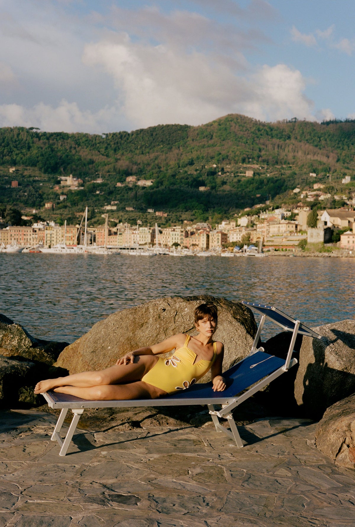 Model reclining in the Flora one-piece in butter yellow and ivory beside the sea, with a coastal town behind her
