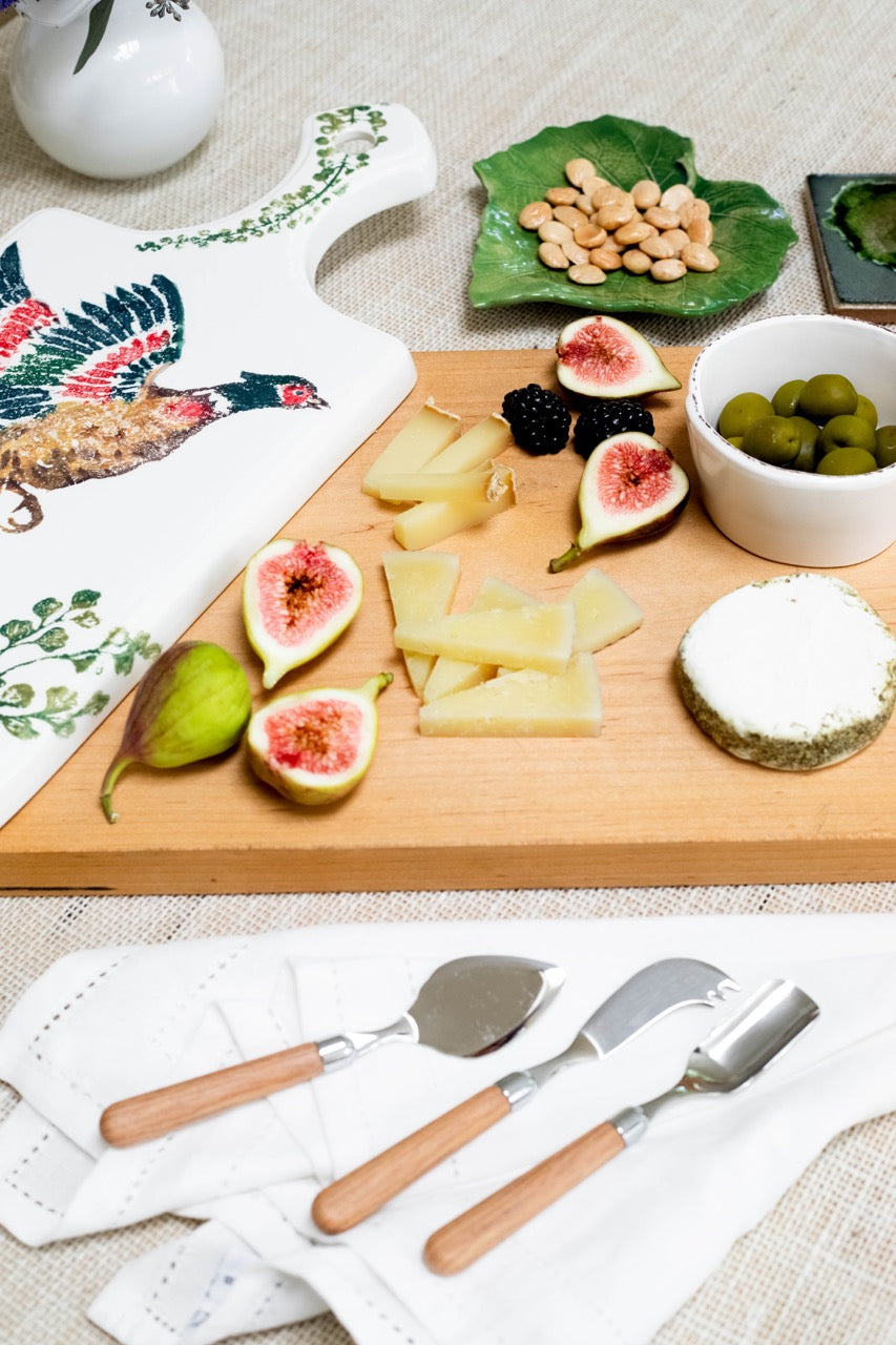 Front view of Albero cheese knife set with elm wood handles and stainless blades on a napkin beside a wooden cheese board