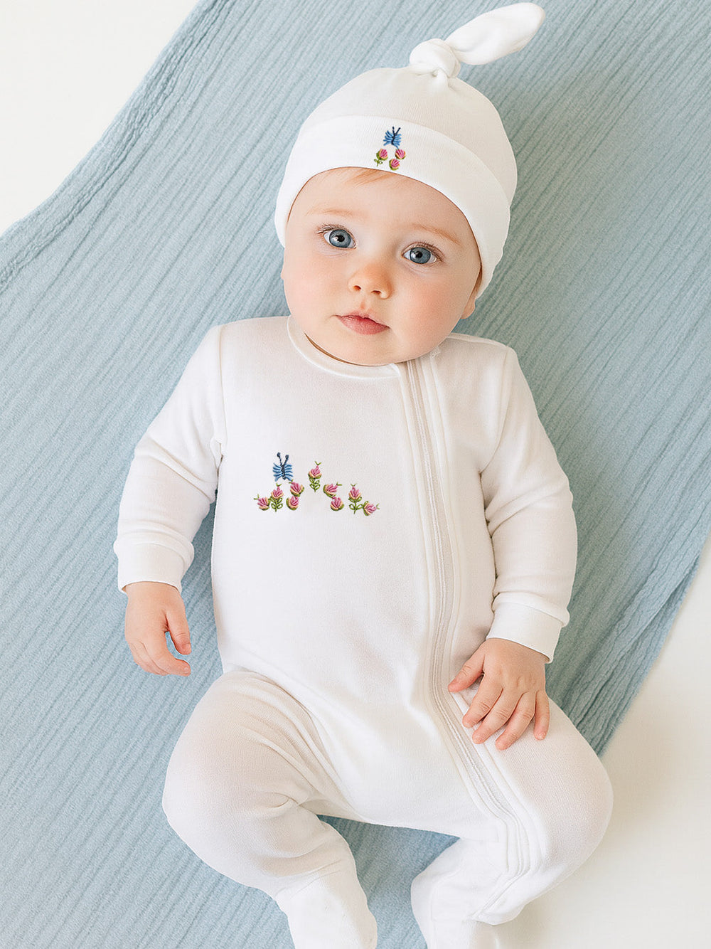 Top view of white combed-cotton baby footie with floral embroidery and zipper, plus matching knotted hat on blue blanket