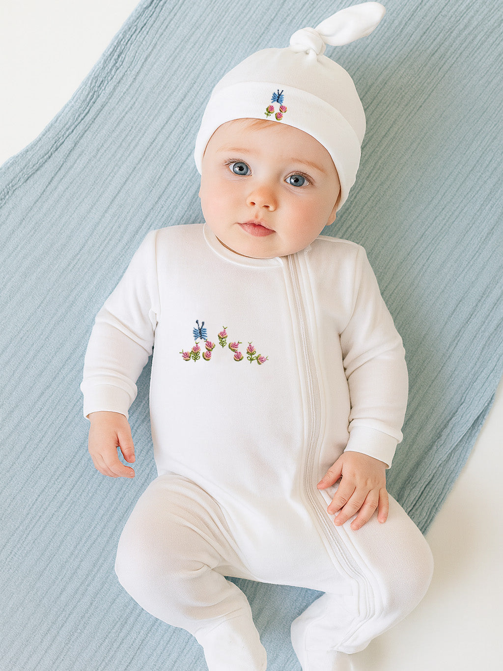 Top view of white combed-cotton baby footie with floral embroidery and zipper, plus matching knotted hat on blue blanket