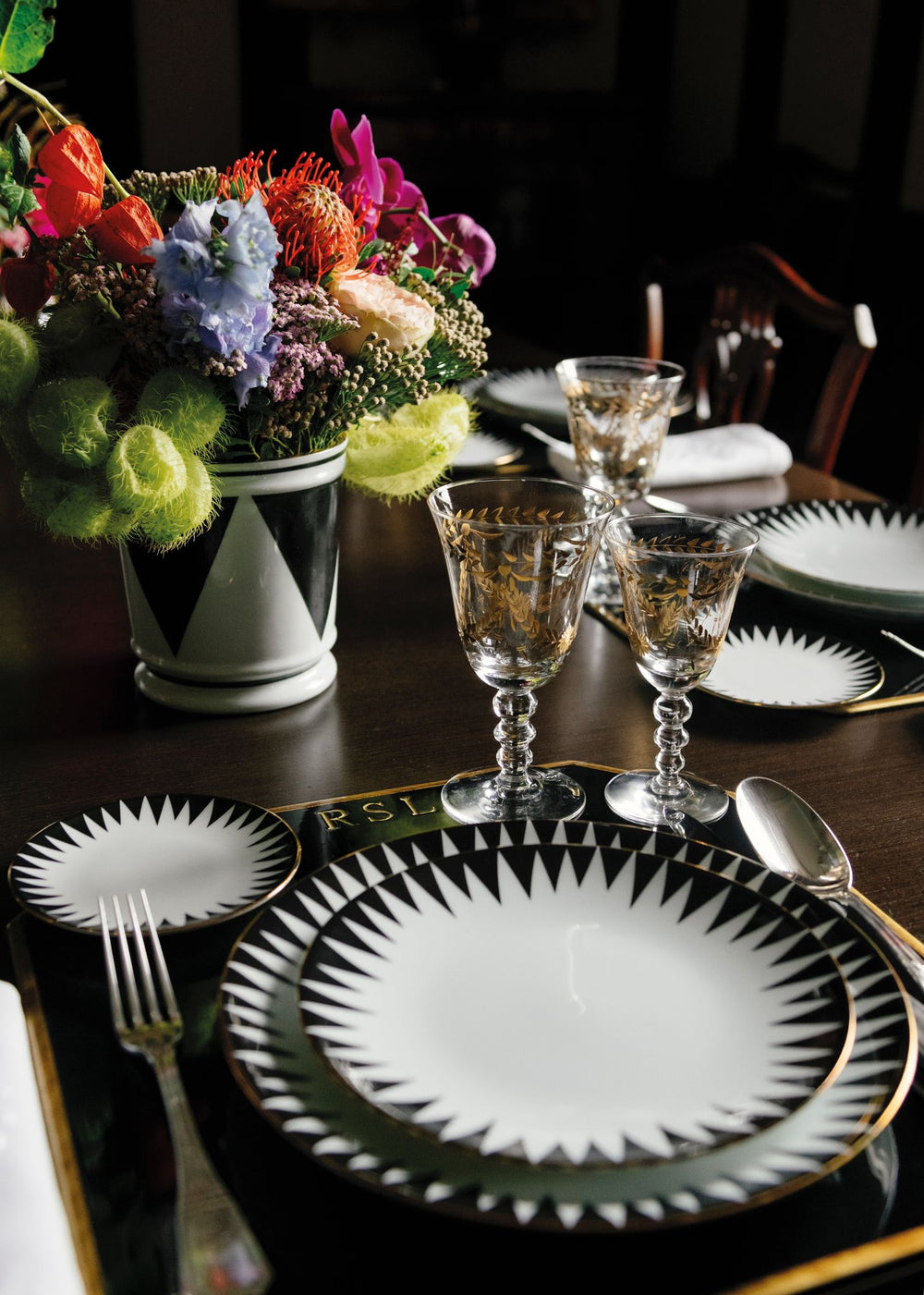 Angled view of Black Punk dinner plate with black starburst border and gold rim on dark wood table with glassware and flowers