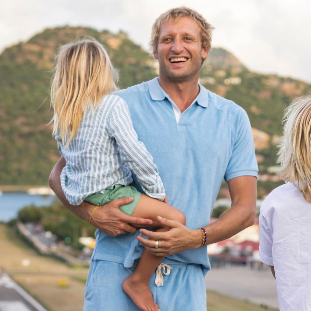 Man wearing Le Marin blue French terry polo smiling while holding a child with coastal hills in the background