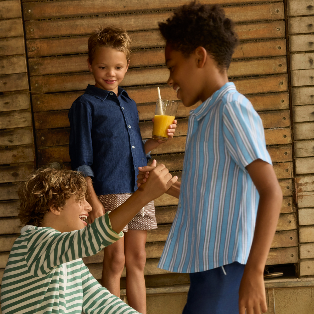 Boys interact outdoors beside a wooden wall, with one wearing a blue striped linen button-down shirt