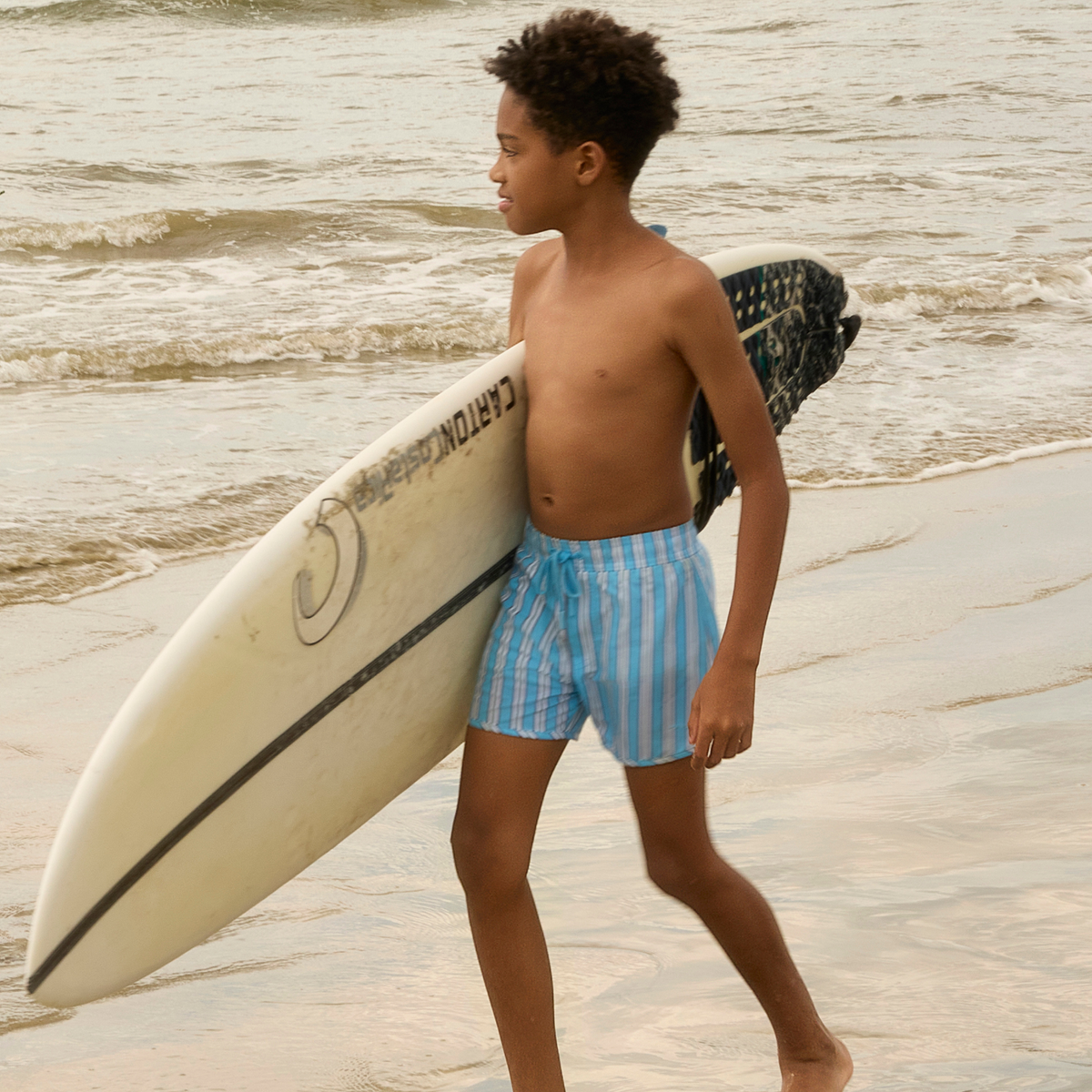 Side view of Boys Isla Blue Stripe Boardie showcasing blue stripes on a boy walking by the beach