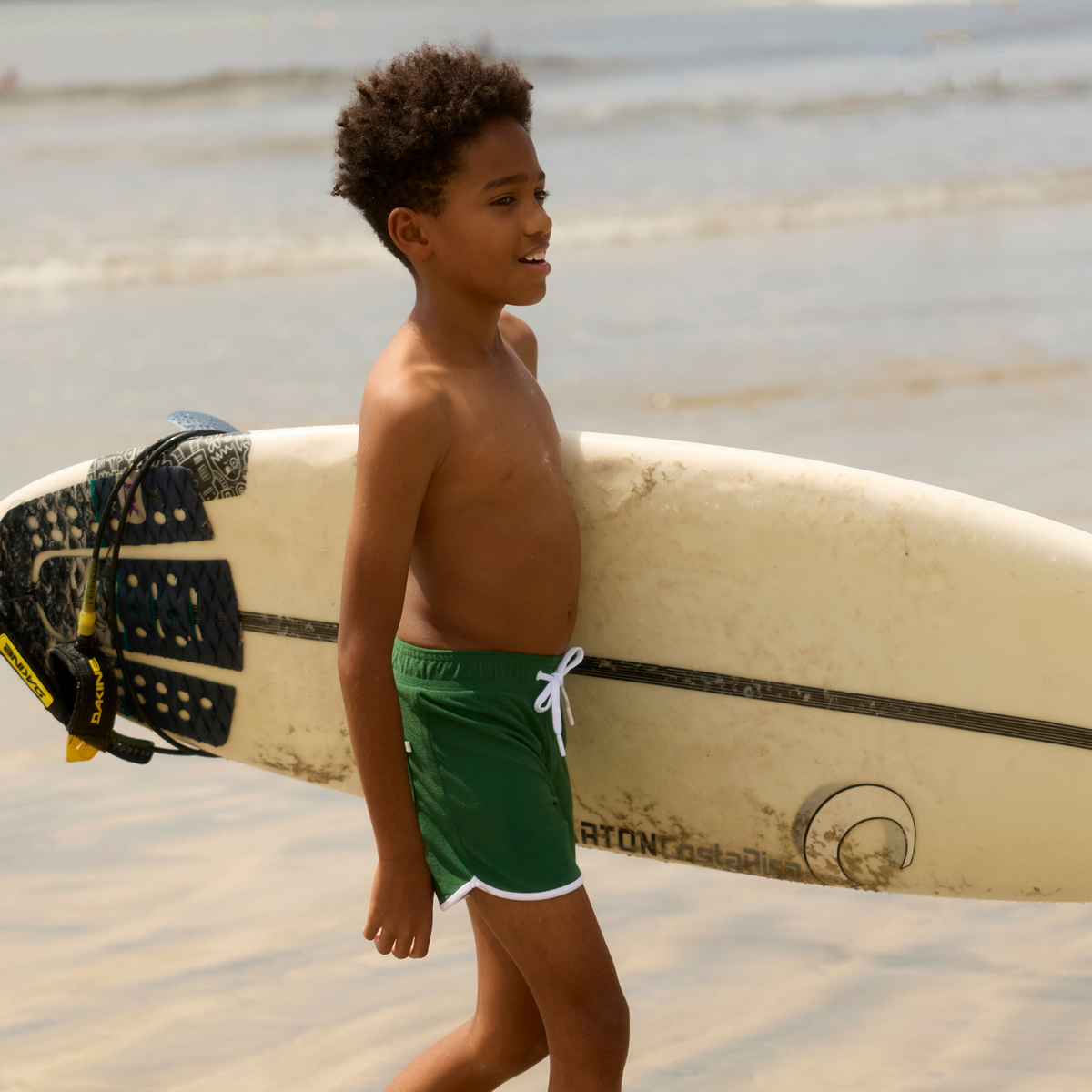 Boy wearing the Palma green terry boardie walks beside a surfboard on the beach