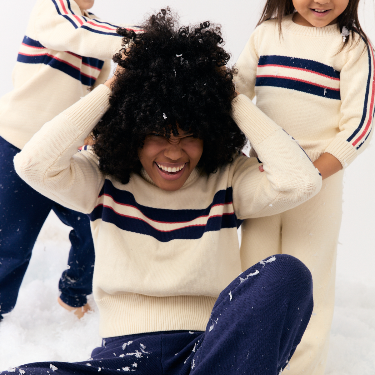 Woman laughing in faux snow wearing a cropped cream knit sweater with navy and red stripes, kids playing behind her