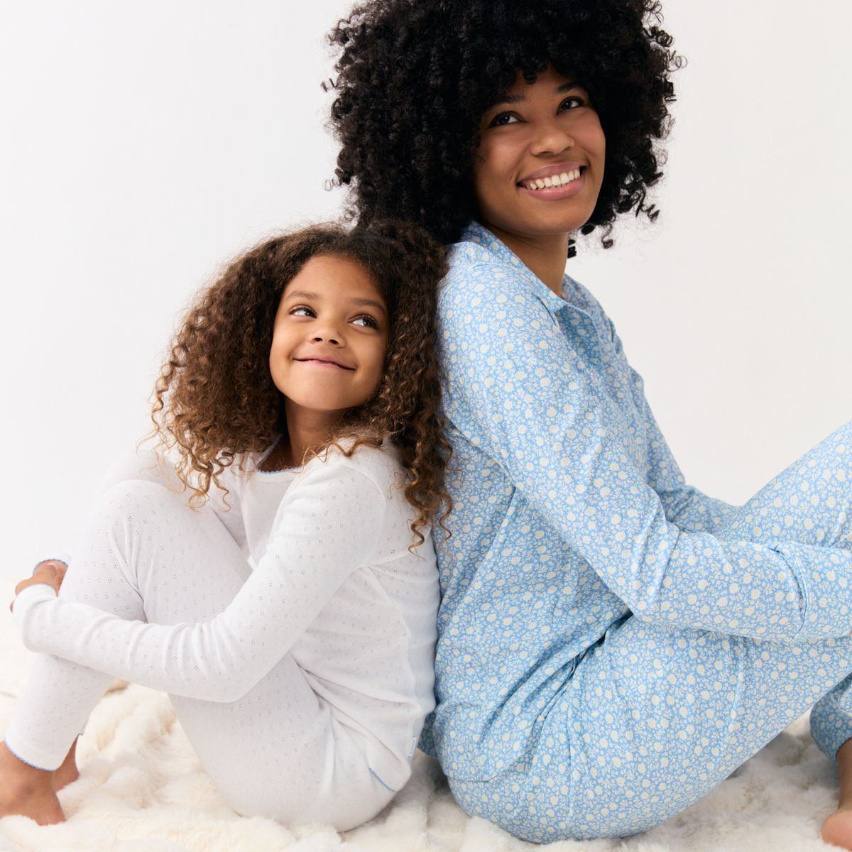 Woman in blue Alpine Fiore collared pajamas sitting back-to-back with a girl in white pajamas on a white rug, both smiling