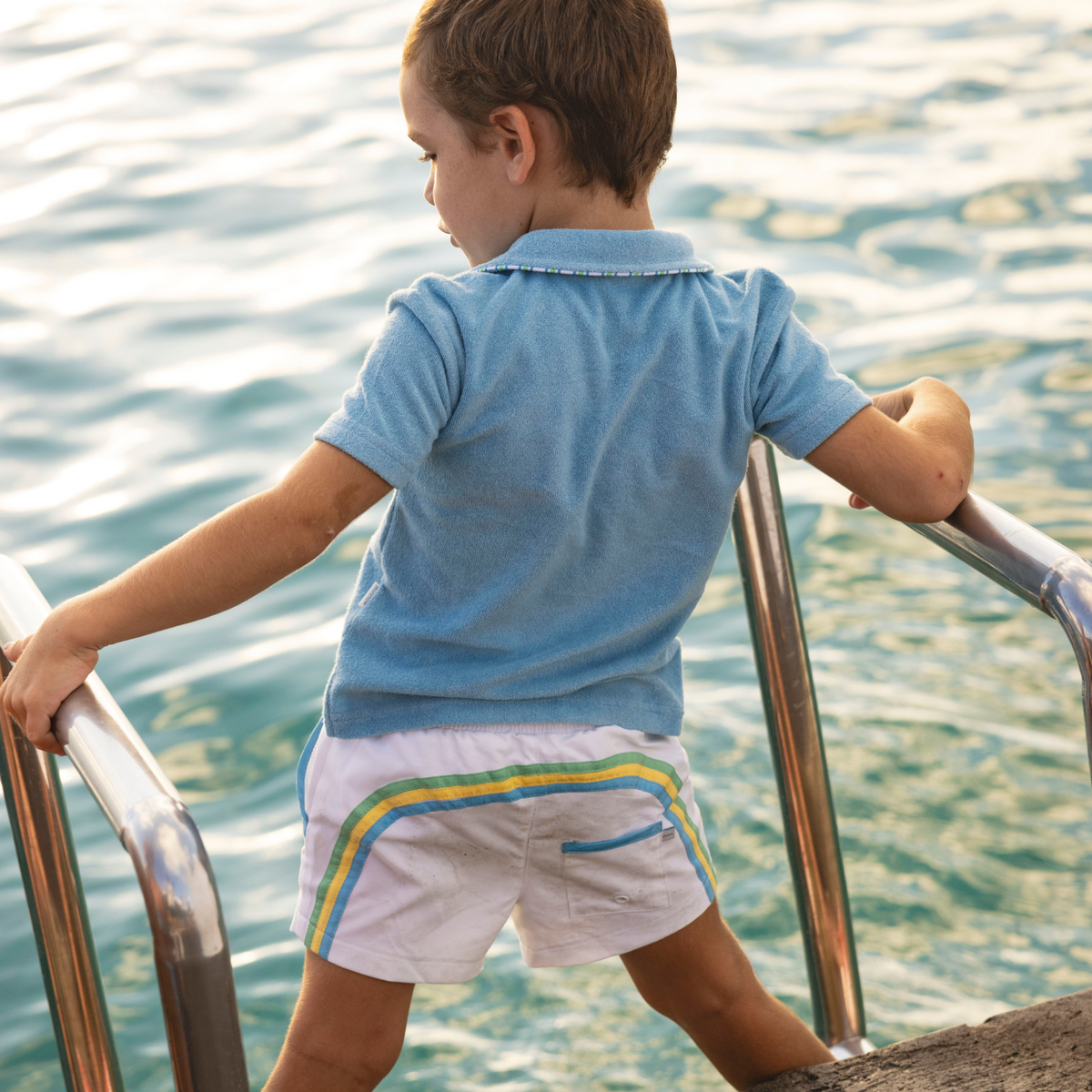 Back view of Boys’ Le Marin blue French terry polo on a child showcasing terry texture and contrast piped collar with sunlit water background