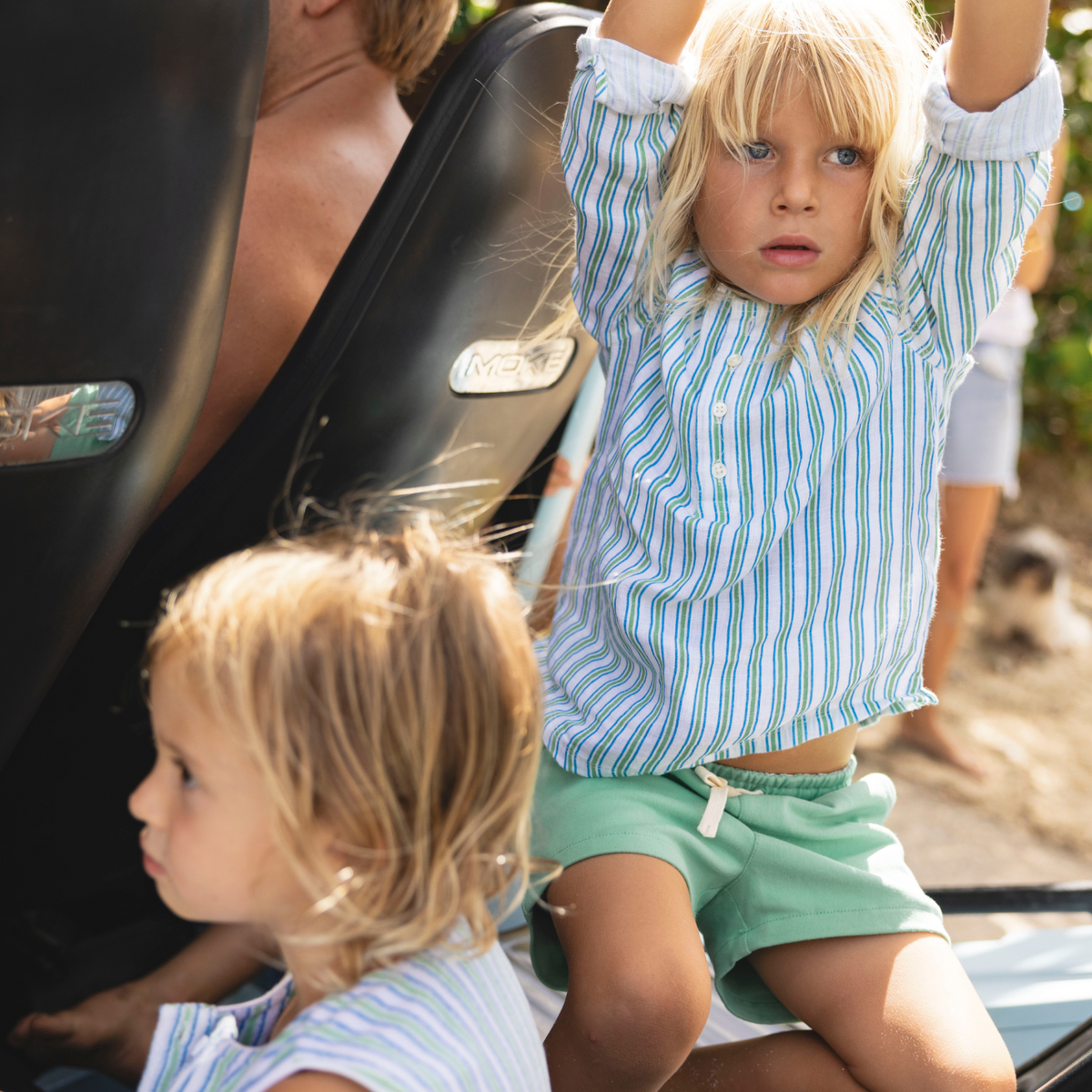 Young boy sitting in a vehicle with his arms raised, wearing green drawstring shorts and a blue striped shirt in a sunny outdoor scene